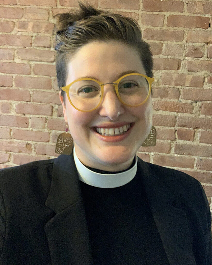 The Reverend Lindsey Briggs is a white woman who appears to be in her/their mid-30s. She has very short hair pulled away from her face. She is wearing round yellow glasses and a priest's collar. She is smiling widely. 